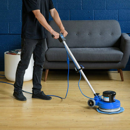Person using a prolux 13  floor polisher on a wooden floor with a gray sofa in the background.